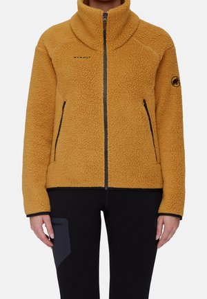 Person iført en sennepsgul fleecejakke med lynlås foran og sorte bukser, stående mod en ensfarvet baggrund.