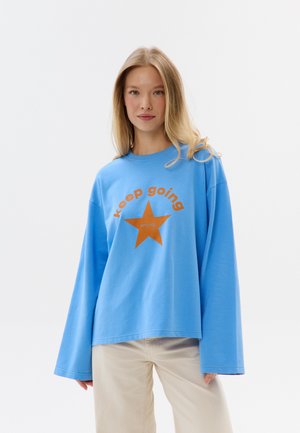Vrouw draagt een losse blauwe blouse met een oranje ster en tekst "keep going", gecombineerd met beige broek, staand tegen een witte achtergrond.