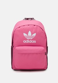 Vaaleanpunainen reppu, jossa on pyöreä muoto, ja se on valmistettu teksturoidusta kankaasta. Sisältää etutaskun ja valkoisen Adidas-logon etupaneelissa.