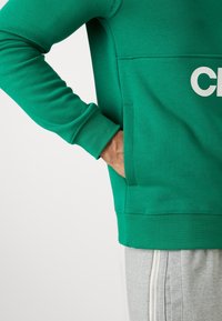 Personne portant un sweat-shirt vert avec une main dans la poche avant et un pantalon de survêtement gris avec une fermeture éclair blanche verticale sur le côté.