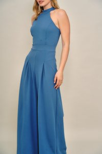 Kék, ujjatlan overall magas nyakkal, testhezálló felsőrésszel és széles, redőzött nadrággal. Sima anyag, szabott kialakítás és hangsúlyos sziluett.