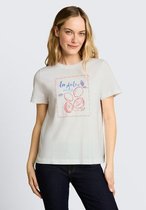 Blondynka ubrana w białą koszulkę z różowym grejpfrutem i napisem „la dolce vita”, do tego ciemnoniebieskie dżinsy, na jasnym tle.