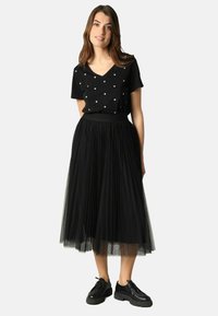 Jupe plissée en tulle noir associée à un haut noir à manches courtes en V orné de petits cœurs. Le mannequin porte des chaussures noires.