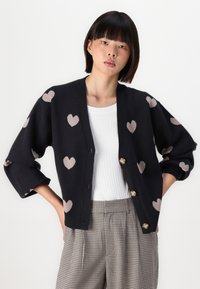Cardigan nero con motivi a cuore marrone chiaro, caratterizzato da una vestibilità comoda, scollatura profonda e due bottoni dorati. Abbinato a una maglietta bianca.
