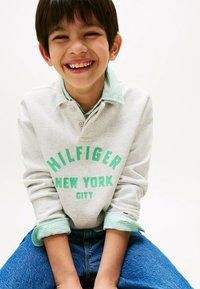 Felpa grigia con testo verde "HILFIGER NEW YORK CITY", sovrapposta a una camicia a righe verde chiaro. Jeans blu e posa informale.