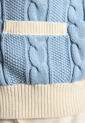 Prim-plan al unui pulover tricotat albastru cu modele în relief tip cablu și punct de semințe, cu margine crem nervurată în partea de jos și o mică secțiune orizontală crem.
