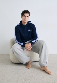 Tamsiai mėlynas džemperis su šviesiai mėlynomis šoninėmis juostelėmis ir Lacoste logotipu, derinamas su pilkais sportiniais kelnių. Minkšta, laisvalaikio medžiaga su laisvu kirpimu.