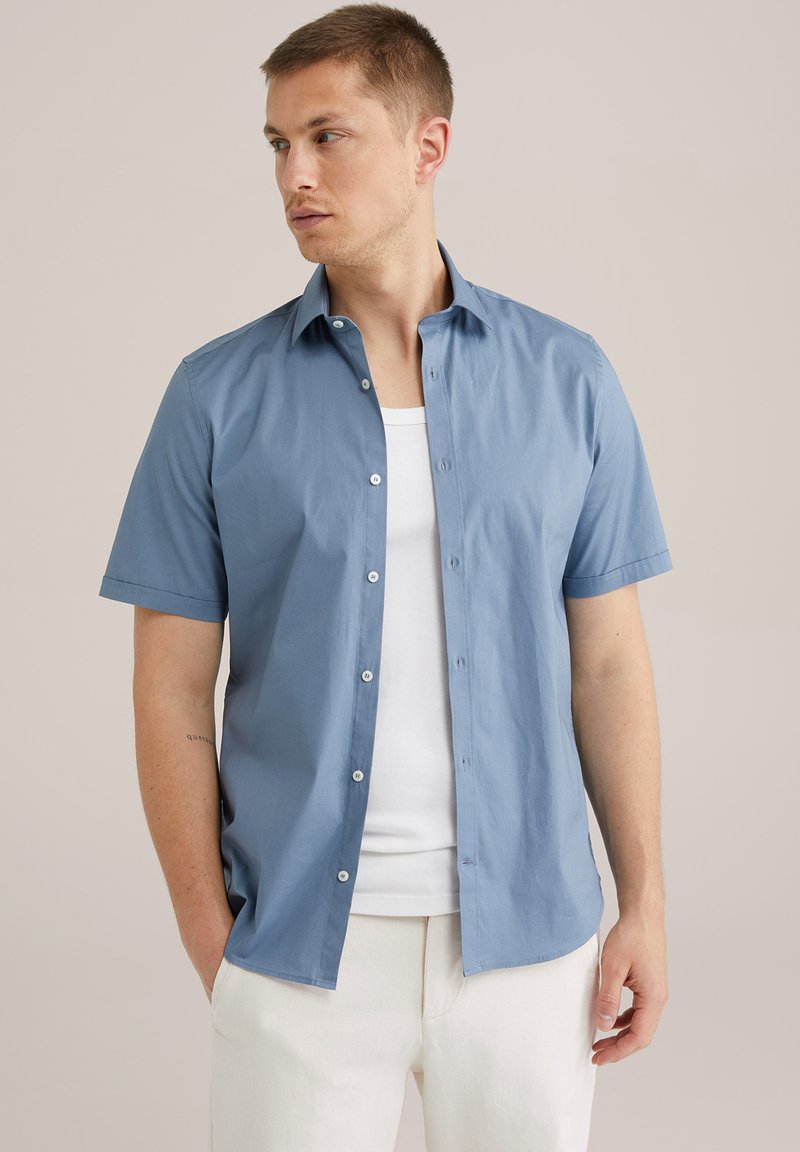 Camisa de manga corta, abotonada, en algodón azul claro, con una textura suave y un collar clásico. Combinada con una camiseta blanca.