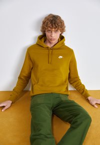 Sweatshirt Nike amarelo mostarda com um bolso frontal e logo branco; combinado com calças verdes. Textura suave, corte relaxado, design casual.