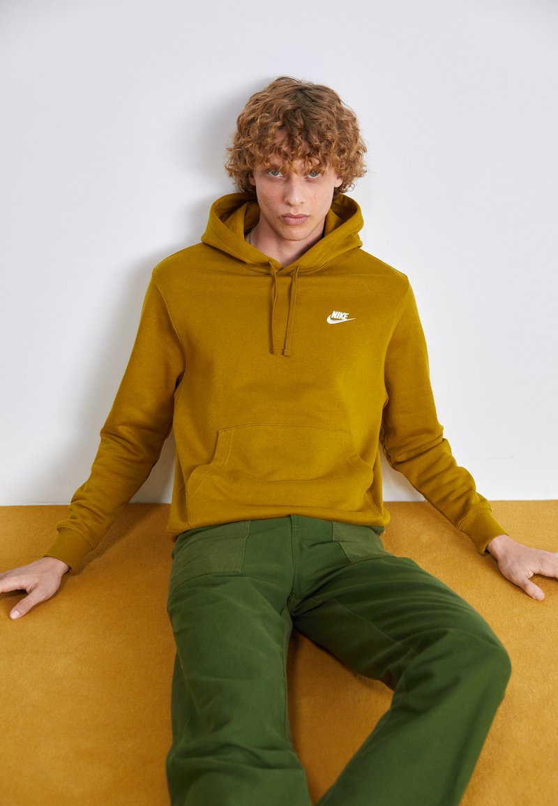 Sweatshirt Nike amarelo mostarda com um bolso frontal e logo branco; combinado com calças verdes. Textura suave, corte relaxado, design casual.