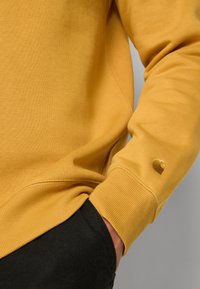 Felpa gialla con polsini a coste, texture liscia, con un piccolo logo sul maniche. Pantaloni neri parzialmente visibili.