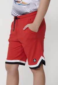 Pantaloni corti rossi con un orlo a righe bianche e nere, cintura elastica e cordino. Presentano tasche laterali e un piccolo logo sulla gamba.