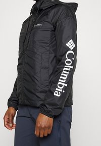 Muž v černé bundě Columbia s kapucí a bílým logem na hrudi a rukávu, v kombinaci s námořnickými kalhotami se zipovou kapsou.