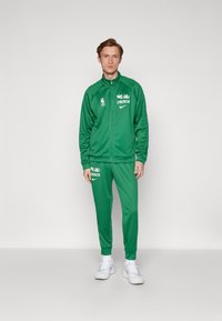 Zeleni trenerak od glatkog materijala, s duksericom na patent, s NBA i Celtics logotipima, odgovarajućim hlačama i bijelim tenisicama.