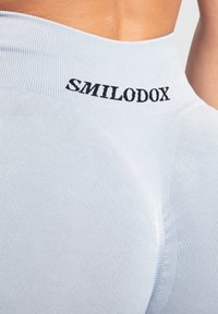 Lichtblauwe stretchstof leggings met een hoge taille. Bevat zwarte tekst "SMILODOX" dicht bij de bovenkant. Glad textuur met subtiele detaillering.