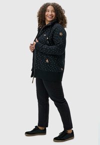 Sudadera negra con un patrón texturizado, con cremallera delantera, capucha ajustable y detalles de madera. Combinada con pantalones negros y zapatos negros.