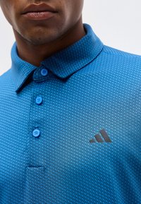 Κοντινό πλάνο ατόμου που φοράει μπλε πικέ πόλο μπλούζα με τρία κουμπιά και σκοτεινό λογότυπο Adidas στο στήθος.