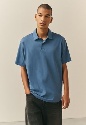 Jonge man met een blauw poloshirt met korte mouwen en een donkere broek, staand met één hand in zijn zak, kijkt naar links tegen een effen achtergrond.