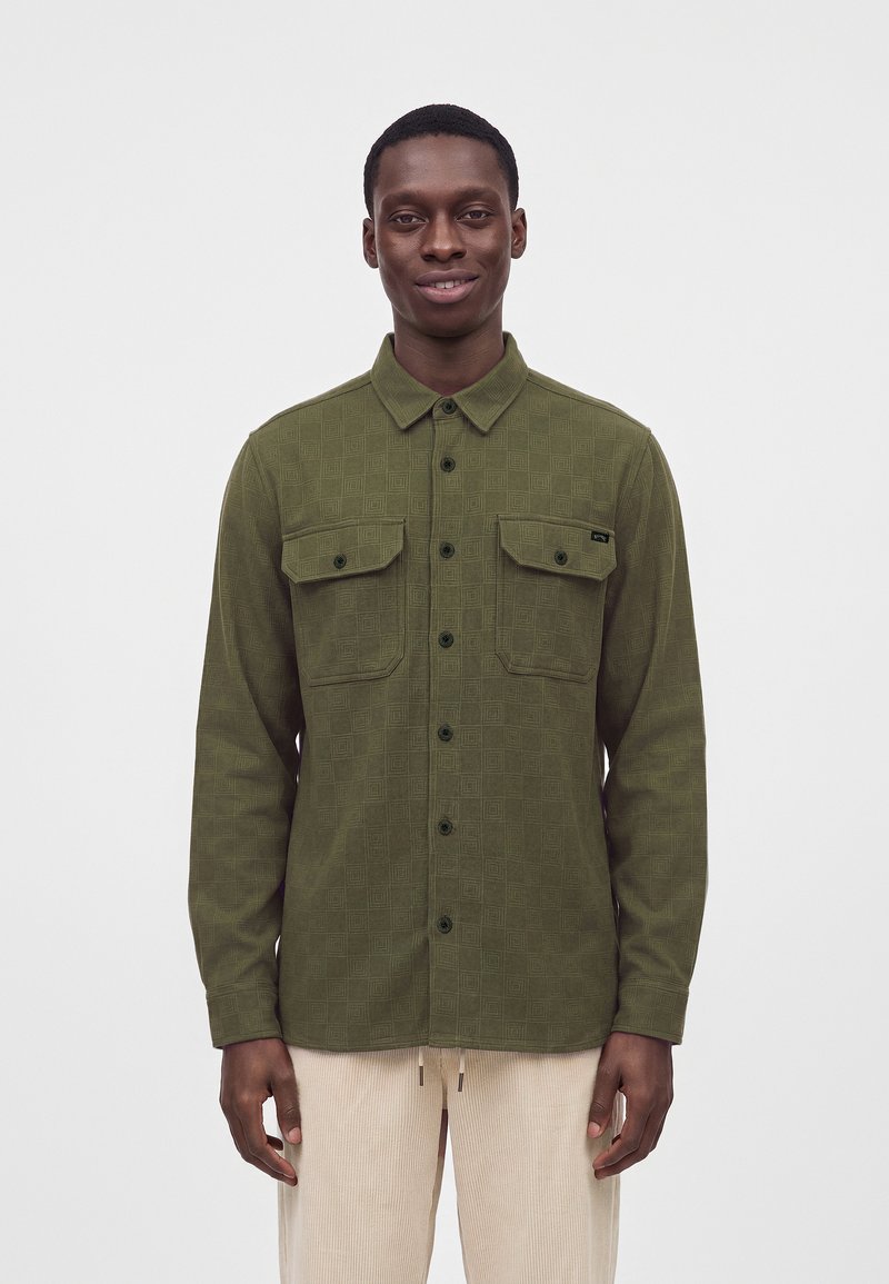 Jeune homme portant une chemise boutonnée vert olive avec deux poches poitrine et un pantalon beige clair, debout devant un fond uni.