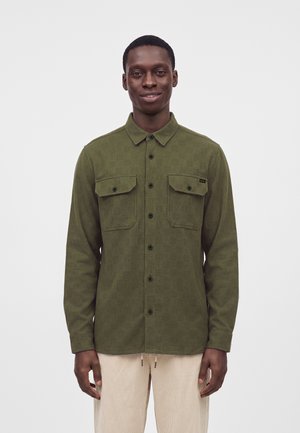 Joven con una camisa verde oliva abotonada con dos bolsillos en el pecho y pantalones beige claros, de pie sobre un fondo liso.
