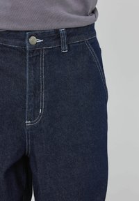 Jeans en denim bleu foncé à coupe droite, dotés de coutures contrastées, d'une fermeture à bouton en métal et de poches avant traditionnelles.