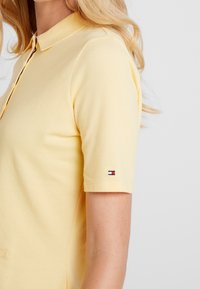 Žluté polo tričko s texturovanou látkou, krátkými rukávy a límečkem. Na rukávu se nachází malé logo s červenými, bílými a modrými akcenty.