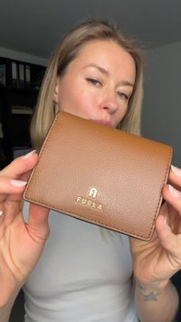 Καφέ δερμάτινο πορτοφόλι με υφασμένο φινίρισμα, που διαθέτει χρυσό λογότυπο μπροστά και ραμμένα άκρα. Συμπαγές ορθογώνιο σχέδιο.