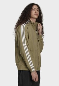 Olivgrön jacka med vita ränder, dragkedjestängning, ståkrage, elastiska ärmslut och två sidofickor. Tillverkad av ett lätt material.