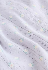 Ljuslila stickat tyg med fina blommönster i pastellfärger, med små knappdetaljer och en strukturerad design.