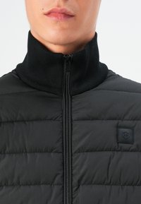 Black puffer jacket with ribbed high neck collar, front zipper, and a textured surface featuring stitched horizontal panels. Diagonal logo patch.