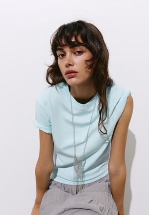 Jeune femme aux cheveux ondulés et foncés portant un haut bleu clair sans manches et un long collier argenté, assise devant un fond blanc uni.