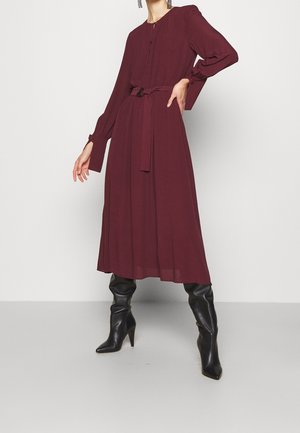 Robe longue à manches longues bordeaux avec une taille cintrée et une ceinture à nouer, présentant une texture plissée. Portée avec des bottes noires à talons aiguilles montant jusqu'aux genoux.