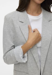 Jasnoszary blazer wykonany z miękkiego materiału; posiada klasyczny kołnierz, dwie przednie kieszenie oraz paski wystające z mankietu.