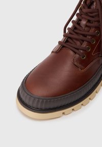 Bottine en cuir marron avec un bout arrondi, semelle en caoutchouc et lacets marron. Présente des accents marron texturés le long de la semelle et des œillets.