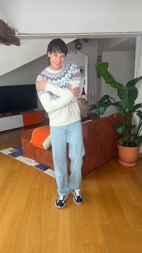 Noor mees mustrilises kampsunis kallistab end ja naeratab elutoas, kus on puidust põrand, oranž diivan, suur potitaime ja telerikapp.