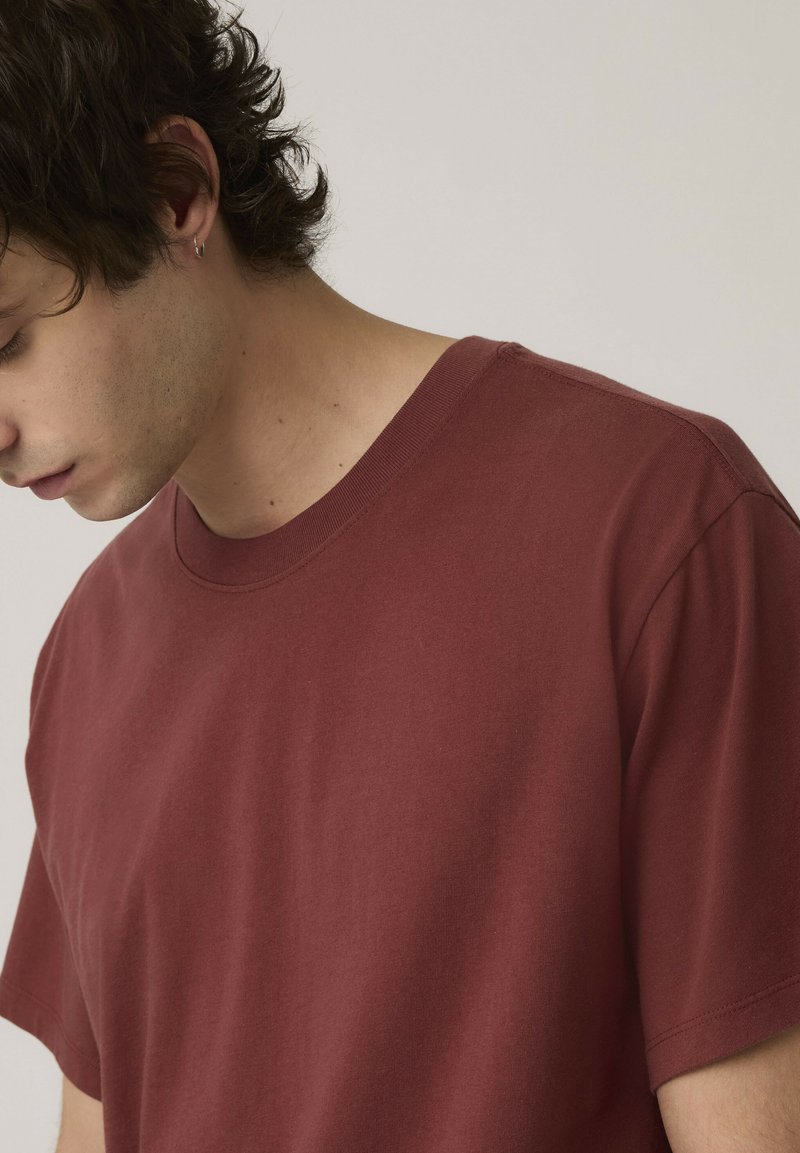 Giovane con capelli corti e ricci e orecchino, che indossa una maglietta bordeaux a maniche corte liscia, guarda verso il basso su uno sfondo neutro.