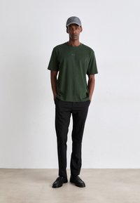 T-shirt en coton vert avec un logo, associé à un pantalon noir. La tenue comprend une casquette grise et des chaussures noires, avec un design épuré et minimal.