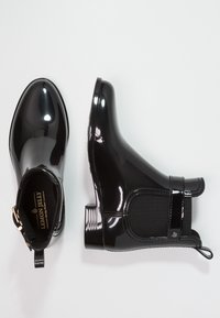Bottes de cheville en vernis noir avec une finition lisse, des panneaux latéraux élastiques et une languette à tirage. Elles possèdent un petit talon carré et des éléments en or discrets.