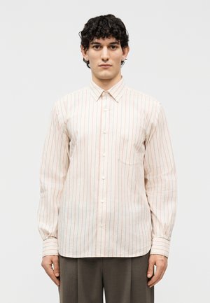 Νεαρός άνδρας με κοντά σγουρά μαλλιά, ντυμένος με ανοιχτόχρωμο πουκάμισο μακρυμάνικο με λεπτές κόκκινες κάθετες ρίγες και σκούρο παντελόνι, στέκεται μπροστά σε μονόχρωμο φόντο.