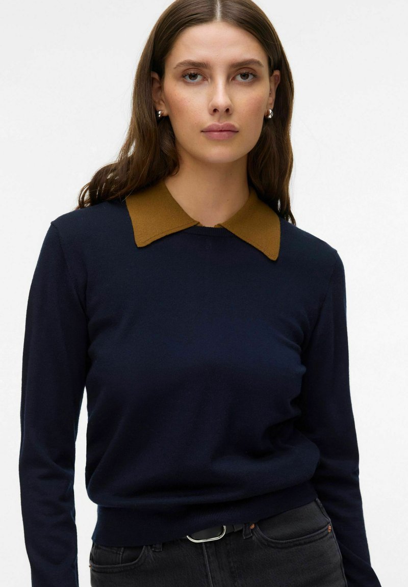 Jeune femme aux longs cheveux foncés portant un pull bleu marine avec un col marron et un jean noir, debout devant un fond blanc uni.
