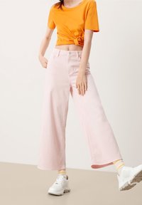 Pantalon large rose clair taille haute, associé à une chemise orange à manches courtes nouée et des baskets blanches avec des accents gris.
