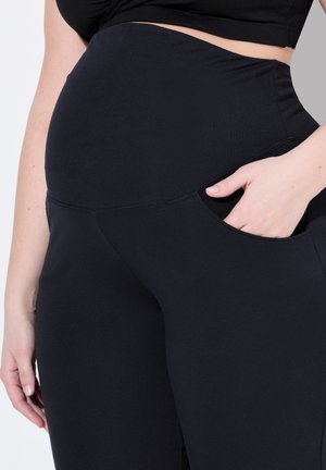 Persona embarazada vistiendo leggings de maternidad negros con una cinturilla alta y con la mano descansando dentro de un bolsillo lateral.