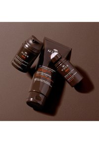 Trois produits de soin NUXE pour hommes : tubes et flacons marron foncé avec des designs épurés, un bouchon pompe, et des étiquettes en texte orange et blanc.