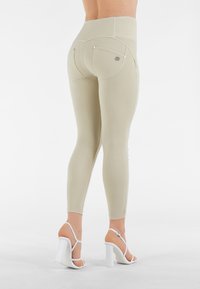 Leggings beige chiaro con vita alta, dotati di tasche posteriori e dettagli in metallo argentato. Texture liscia e design aderente. Tacchi bianchi.