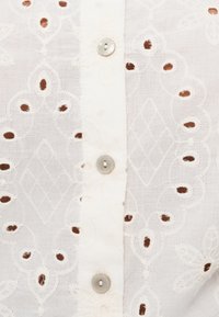 Λευκό κεντητό πουκάμισο με περίπλοκα λουλουδένια και ρόμβου μοτίβα, με μικρές λεπτομέρειες κοψίματος και ασημένια κουμπιά κατά μήκος του μπροστινού μέρους.