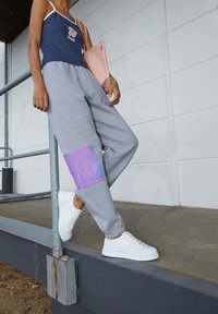 Pantalones deportivos grises con un parche gráfico colorido, combinados con una camiseta sin mangas azul marino con un logo y zapatillas blancas. Se sostiene una bolsa rosa.