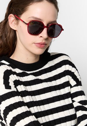 Giovane donna che indossa occhiali da sole esagonali rosso scuro e un maglione a righe nere e bianche con capelli castani lunghi.