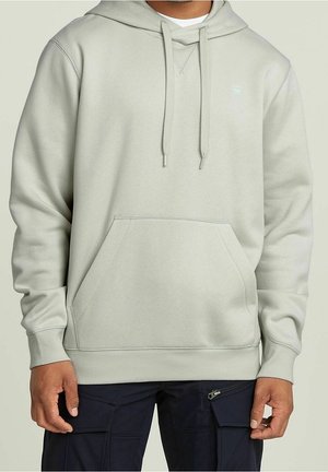 Jasnoszara bluza z kapturem z przednią kieszenią typu kangur, noszona z ciemnymi spodniami cargo, częściowo widoczne dłonie spoczywające po bokach.