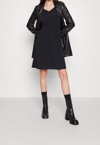 Musta mekko, jossa on istuva yläosa ja avara helma, yhdistettynä mustaan nahkatakkia ja nilkkureihin, joissa on neliökorot.