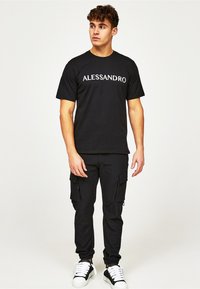 Musta puuvillane t-särk, millel on valgete tähtedega trükitud "ALESSANDRO". Kombineeritud mustade cargo-pükstega, millel on tõmblukuga taskud. Kannab musti ja valgeid tossusid.